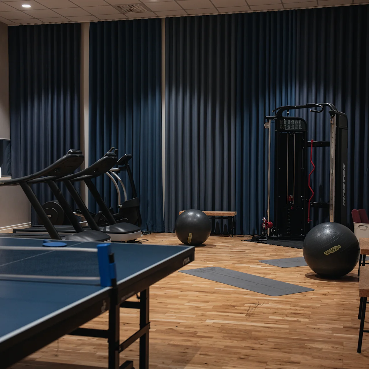 Pingisbord står i ett fitnessrum med trägolv, med löpband, träningsbollar och gymutrustning bakom; Blå gardiner täcker en vägg. Bänkar och en TV är på sidorna.
