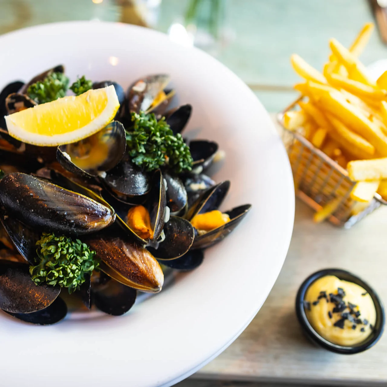 Muslinger pyntet med citron og persille, serveret i en hvid skål, ledsaget af pommes frites i en kurv og en lille skål med cremet sauce på et træbord.
