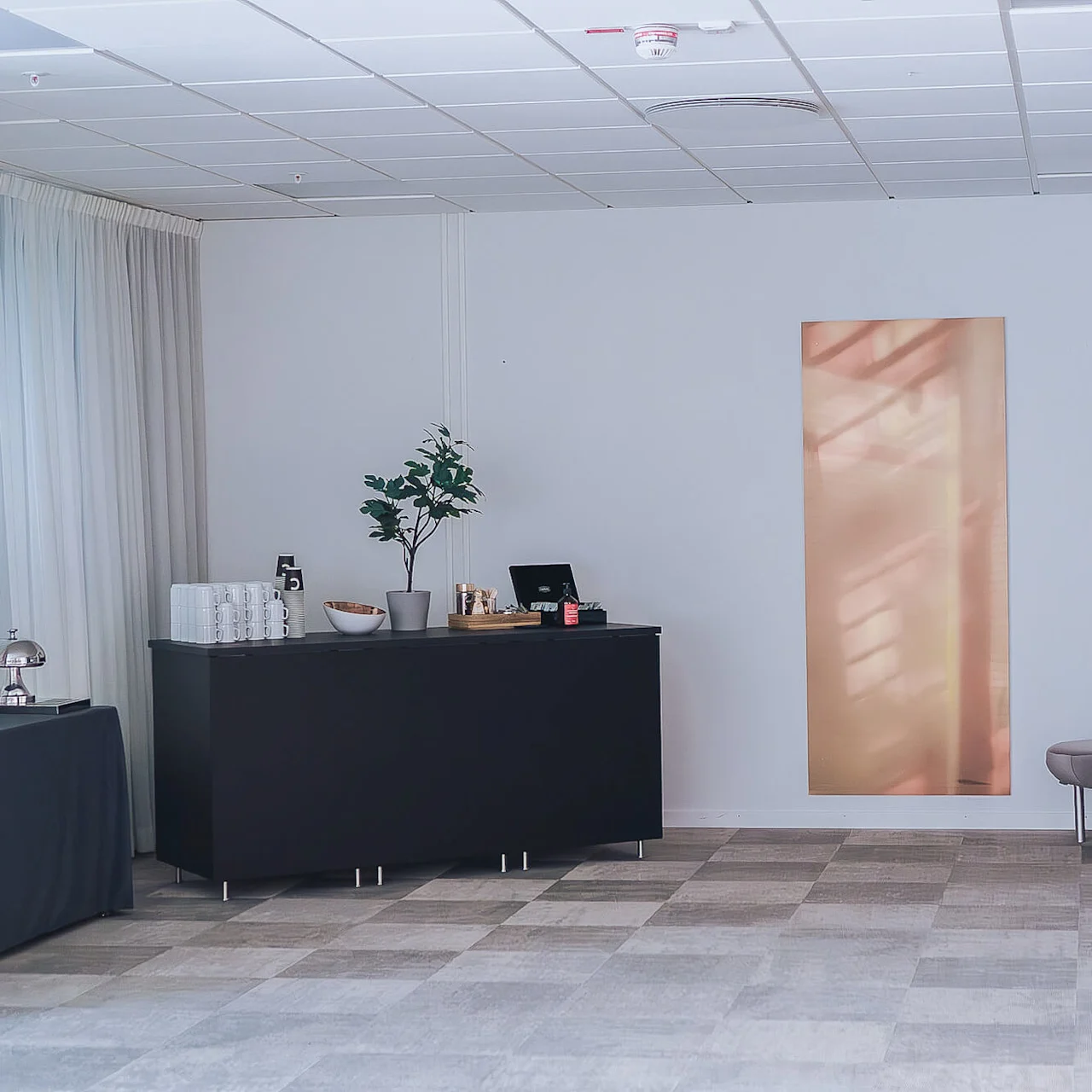 Et bord har stablede kopper, en potteplante, en klokke, og nogle skåle i et minimalistisk rum med grå vægge, en bænk og blød belysning.