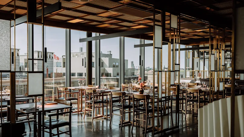 A dining area with wooden tables and chairs sits empty, surrounded by modern glass walls, overlooking a cityscape under clear blue skies.