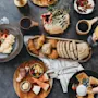 Tallerkener med morgenmadsmad, herunder bacon, æg, croissanter og frugter, er arrangeret på et mørkt bord med kaffe- og juiceglas, der skaber en brunchstemning. Hænder holder tallerkener og glas.