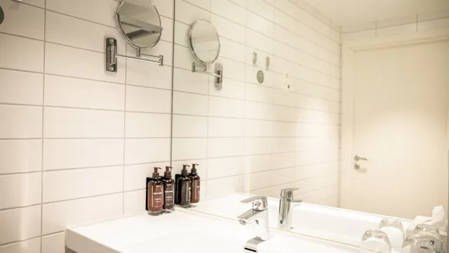 Moderne bad med dobbeltvask, hvite flisvegger og et stort speil. Såpedispensere og glass plassert på benken. En lukket dør er synlig i bakgrunnen.