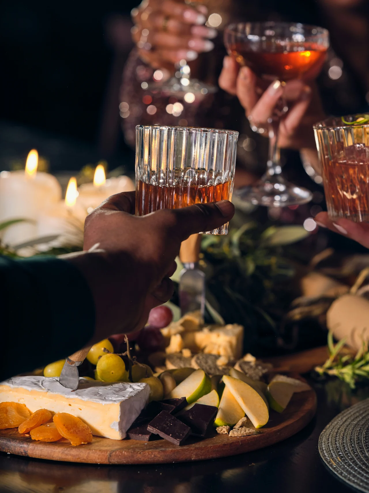 Glasögon klingar i en skål; fyra personer firar runt ett bord med levande ljus och en mängd ost, frukt och garneringar, vilket skapar en varm, festlig atmosfär.