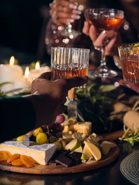 Glasögon klingar i en skål; fyra personer firar runt ett bord med levande ljus och en mängd ost, frukt och garneringar, vilket skapar en varm, festlig atmosfär.