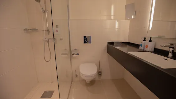 Et moderne bad har en dusj med glassvegger, veggmontert toalett og mørk benkeplate med servant. Såpe dispensere og speil er på høyre side.