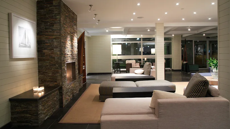 Modern, cozy living room features a stone fireplace, adjacent couch seating, soft lighting from ceiling spotlights and lamps, with a large window providing night views. Wall art depicts a sailboat.