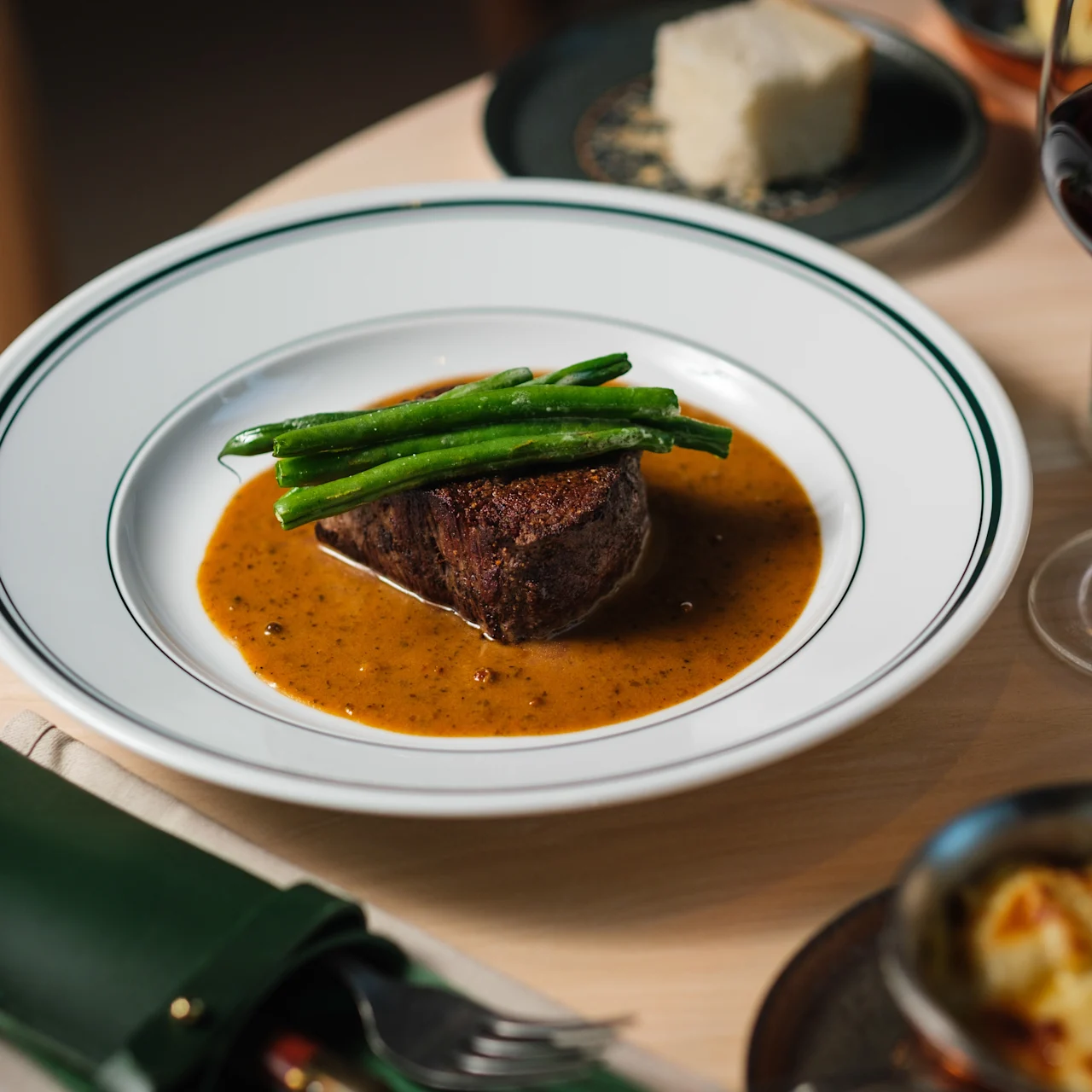 En biff toppas med gröna bönor och serveras i en brun sås på en vit tallrik, omgiven av ett glas rödvin, bröd och bestick på ett träbord.