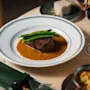 En biff toppas med gröna bönor och serveras i en brun sås på en vit tallrik, omgiven av ett glas rödvin, bröd och bestick på ett träbord.