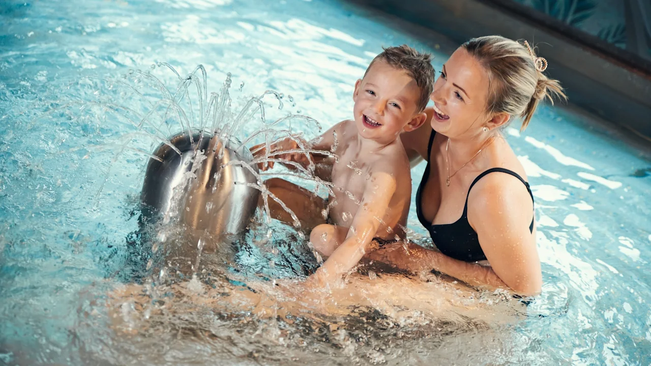 A child and an adult play joyfully in a shallow pool, splashing water around a fountain-like feature. The background shows poolside tiles and a light, watery environment.