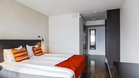 Et koselig hotellrom med en stor seng, livlige puter og et lyst teppe. En gang leder til et speil som reflekterer mykt lys, og skaper en innbydende atmosfære.