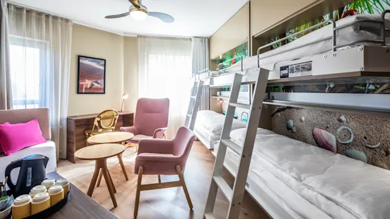 Bunk beds are aligned against a wall in a bright room with wooden floors. Nearby, pink chairs and a small round table sit near a window. Amenities include a desk and a tray with a kettle.