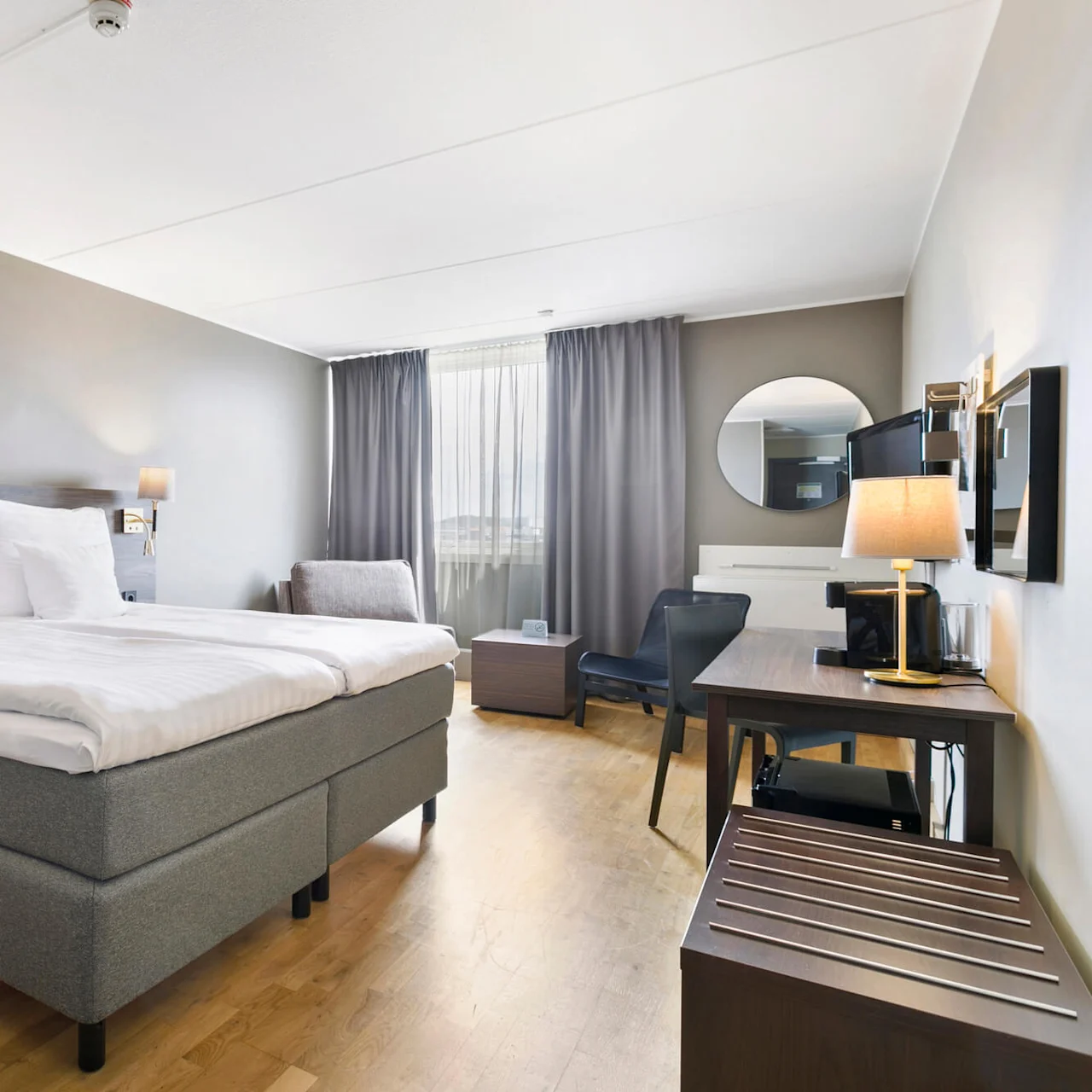 Et pænt og velordnet hotelværelse har en dobbeltseng med hvidt linned, et træbord med en lampe og et fladskærms-TV. Gardiner dækker et vindue, med neutrale farver på vægge og gulv. 