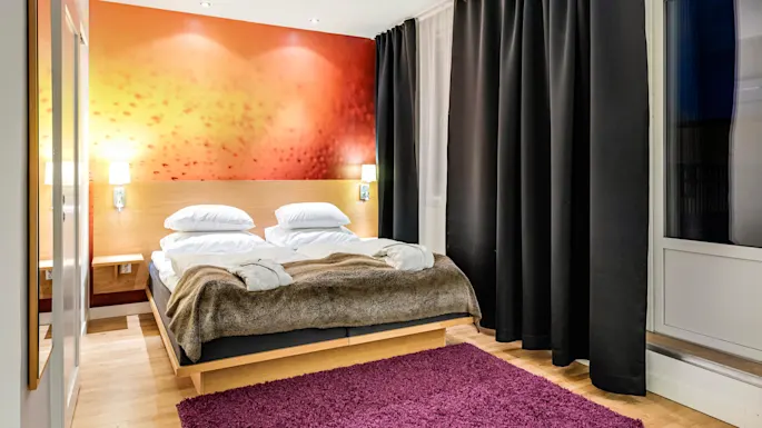 En seng med hvite puter og et brunt teppe står mot en oransje aksentvegg, flankert av nattbordslamper, ved siden av et vindu med mørke gardiner.