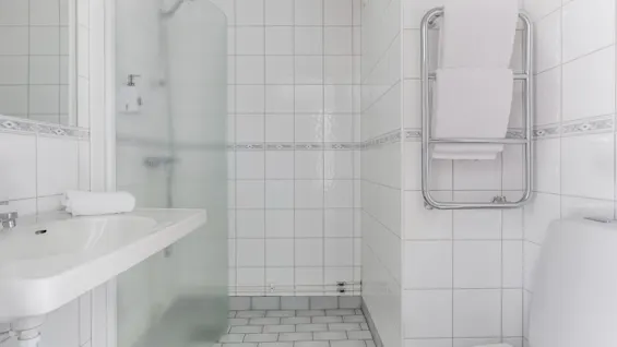 A clean, modern bathroom with white tiles, a sink, a shower, a toilet, and fresh towels on a heated rack.