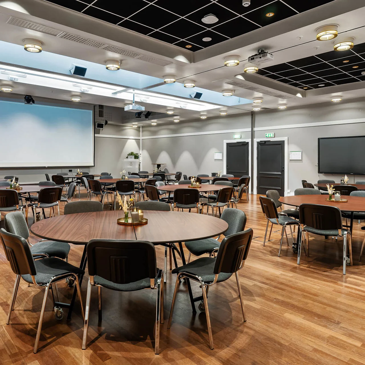 Runde borde med stole arrangeret i et rummeligt konferencelokale; en stor skærm, projektor og moderne belysning fremhæver det professionelle miljø. Grønne planter og minimalistisk dekoration tilføjer subtile detaljer.