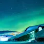 Snedækkede bygninger lyser op under et nordlys på en vinternat.