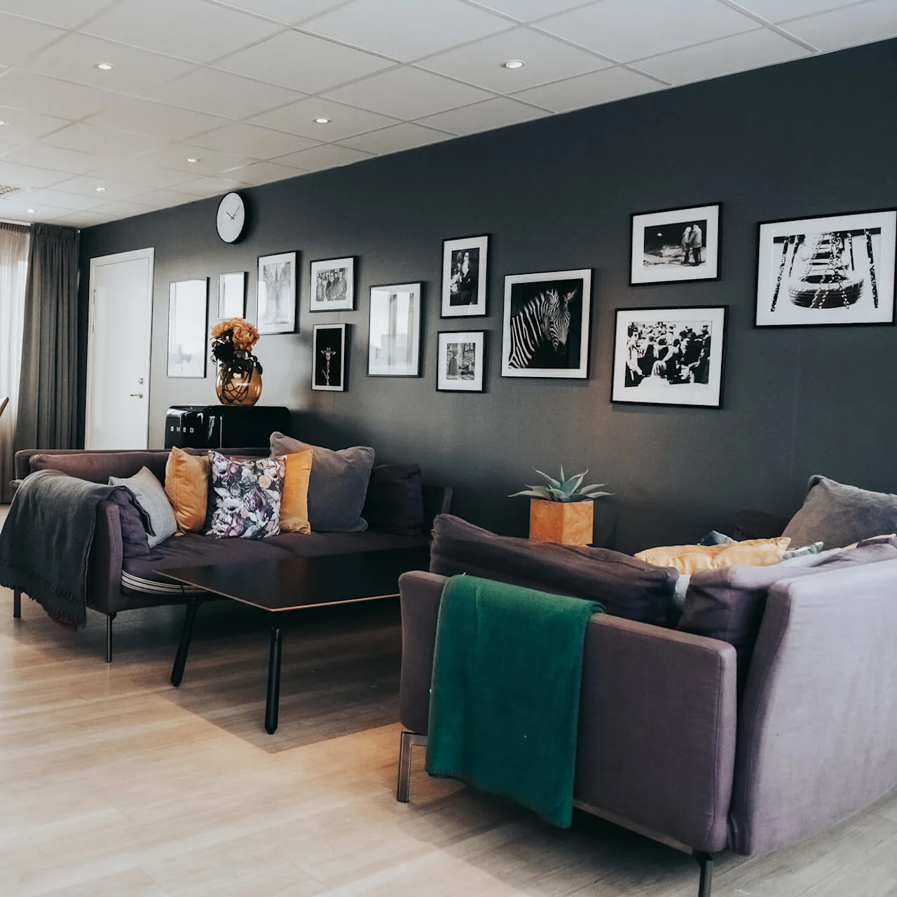 A modern living room features a gray sofa with colorful cushions, facing a wall adorned with framed photos. A dining table and chairs sit nearby under large windows.