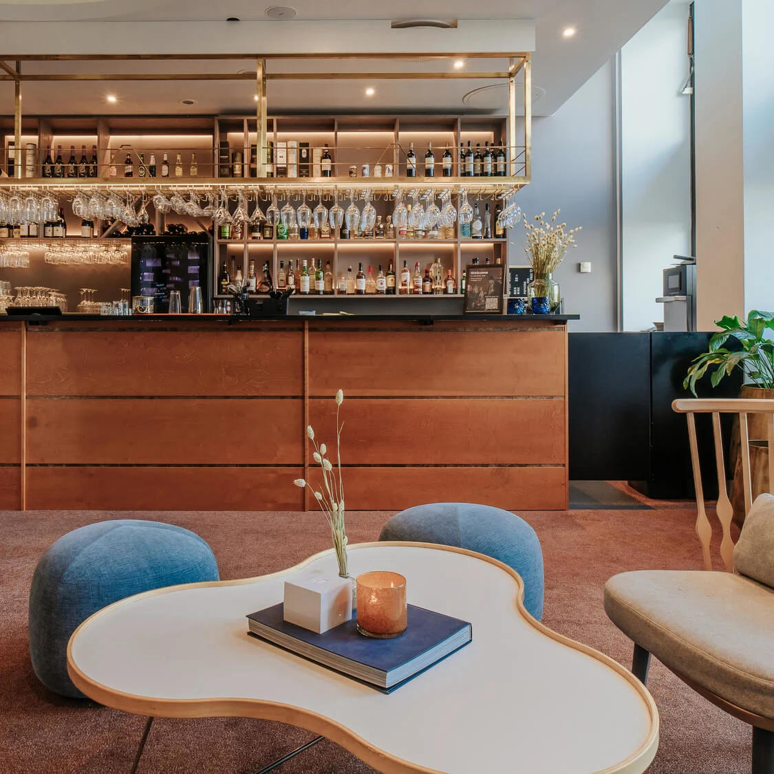 Bar shelves displaying various bottles and glasses above a wooden counter in a modern lounge with blue chairs and a uniquely shaped white table.