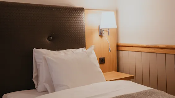 A comfortable hotel bed with white linens, a brown headboard, and a warm bedside lamp, inviting relaxation.