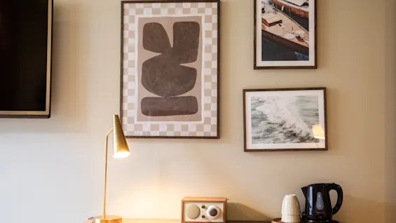 Ett skrivbord har en lampa, radio och vattenkokare, med beige väggar och tre inramade konstverk. Ovanför är en TV monterad på vänster sida.