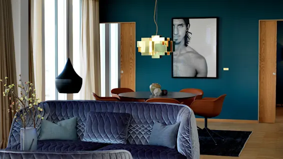 En quiltad blå soffa med kuddar står mot en modern teal vägg prydd med ett stort porträtt. Ovanför hänger en geometrisk lampa över ett matbord med orange stolar.