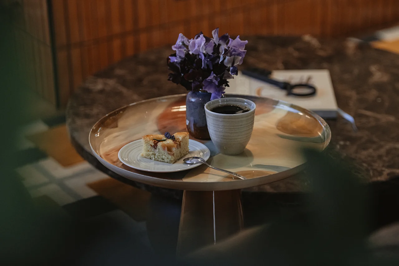 En härlig fikastund med en bit kaka, en varm kopp kaffe och en liten vas med lila blommor på en bricka.