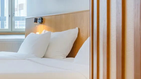 A neatly made bed with white linen, seen through a partially open door, in a modern room with a window.