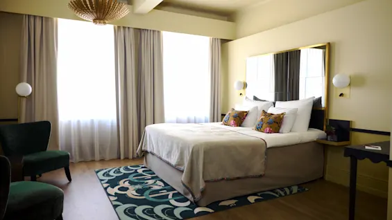 A bed with pillows sits against a wall in a sunlit room with curtains, a rug, and a green chair. A chandelier hangs from the ceiling.