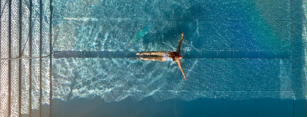 Aerial view of a person swimming in a clear blue pool with steps, surrounded by tiling.