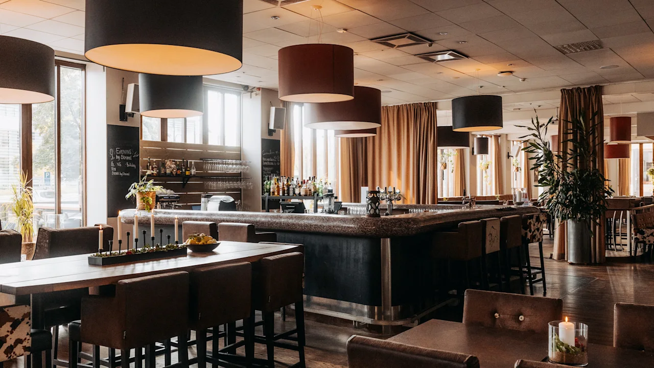 Tyylikäs hotellin baari- ja ravintola-alue, jossa on pitkä baari, mukavat istuimet ja suuret ikkunat. Teksti Evening By Strand.