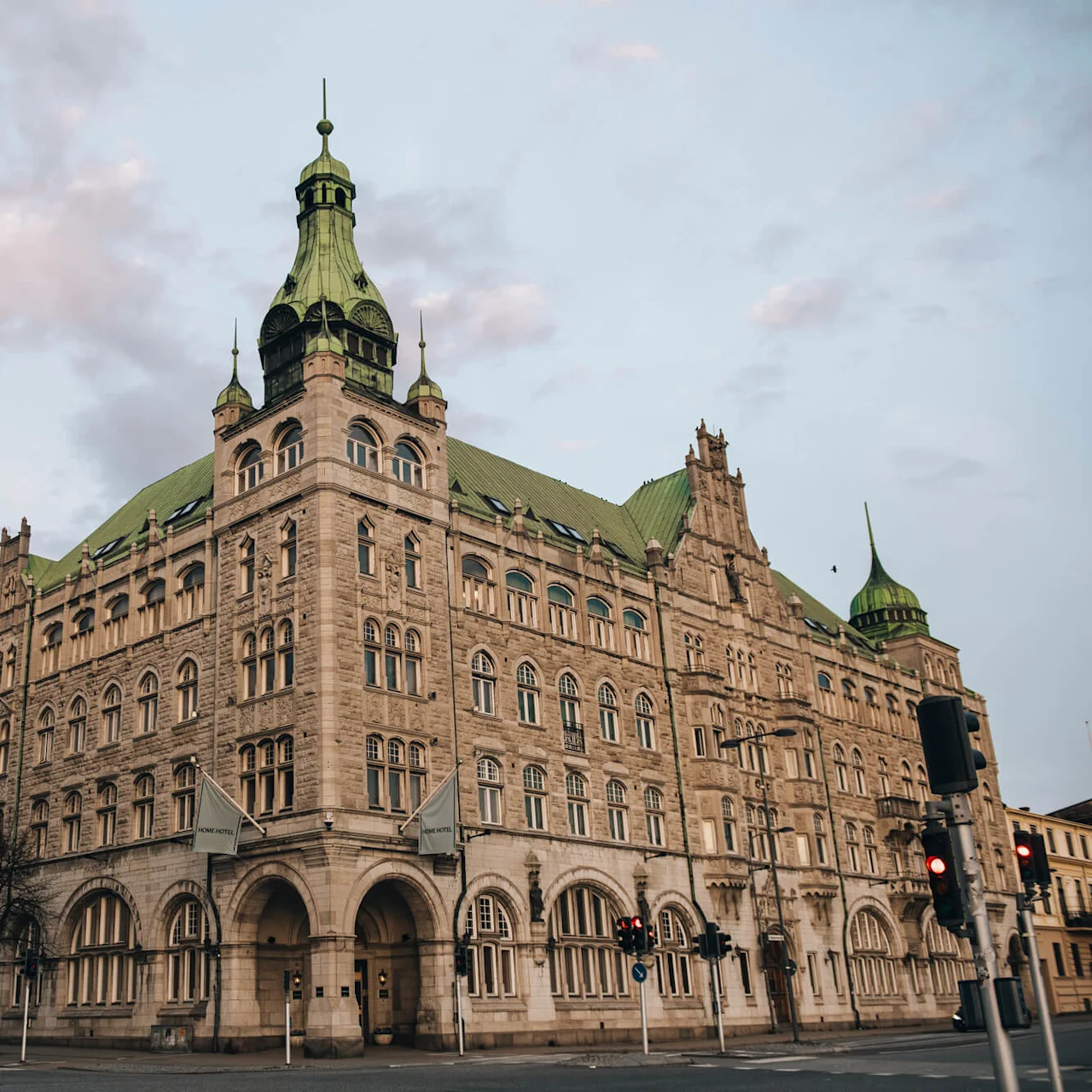 A large, ornate stone building with a green roof stands on a street corner. Surrounding it are other historic buildings and traffic lights. The sky is cloudy, casting a warm light.