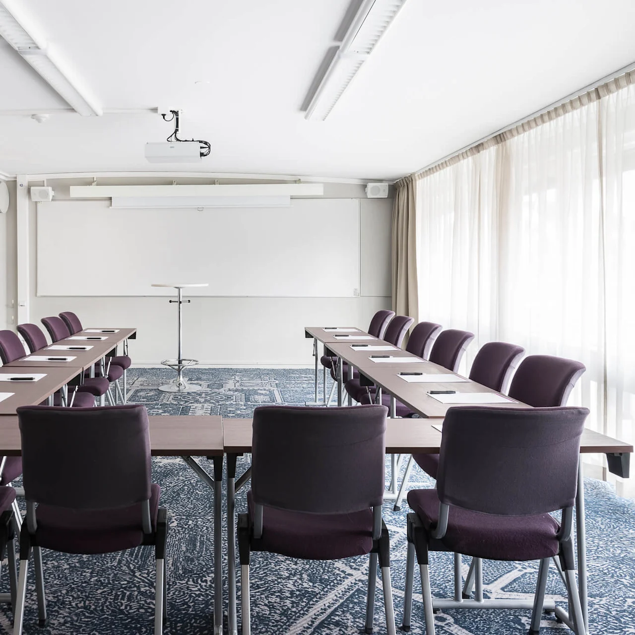 Ett konferensrum med prydligt arrangerade stolar och bord i en U-form, vänd mot en whiteboard, under ljusa taklampor och omgiven av tunna gardiner.
