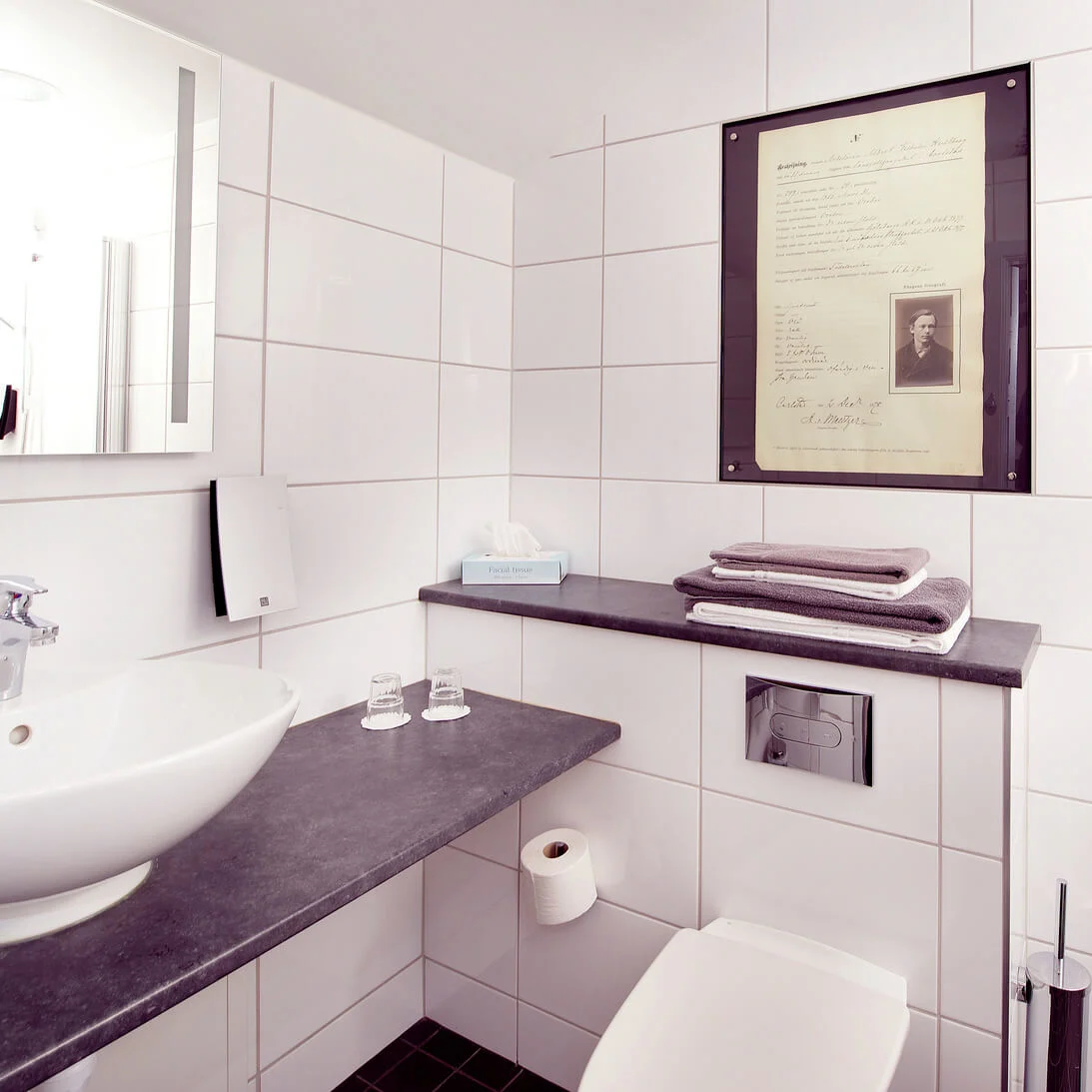 A modern bathroom with white tiles, featuring a sink, mirror, framed document; towels on a shelf add a touch of comfort.