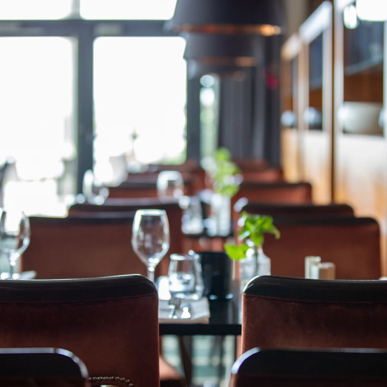 Bord dekket med glass og tallerkener venter på gjester i en mykt opplyst restaurant, med brune stoler og dekorative planter, med store vinduer som gir naturlig lys i bakgrunnen.