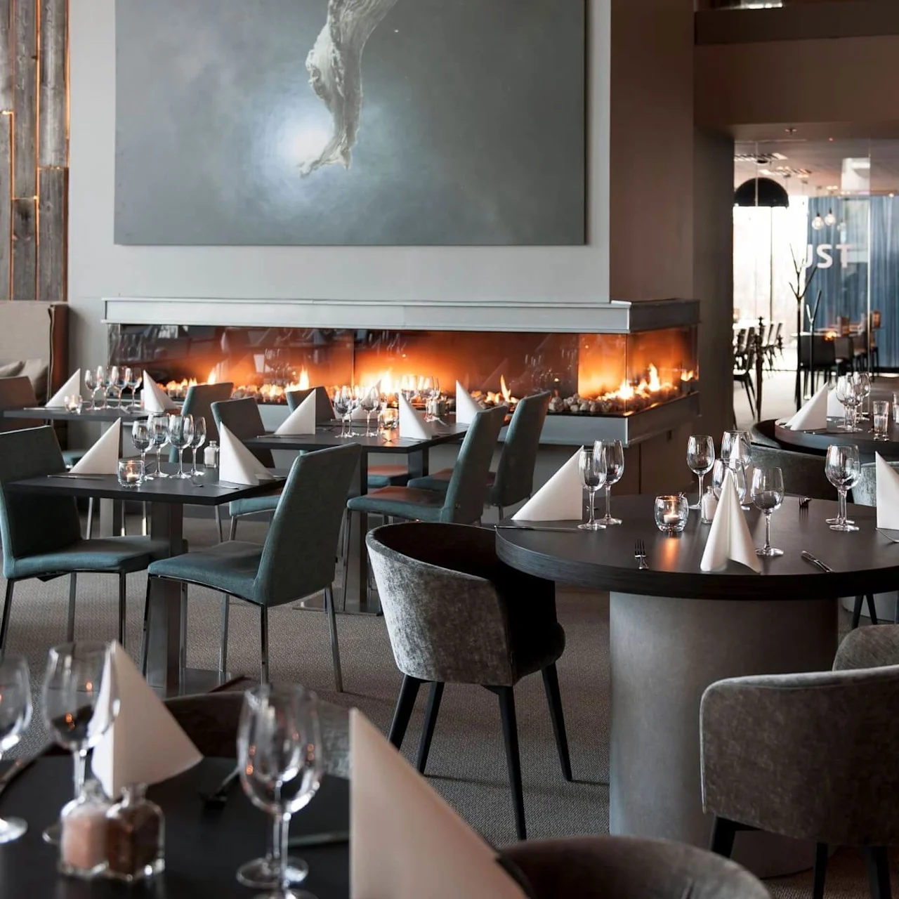 Elegante restaurantbord dekket med glass og brettede servietter, plassert ved en moderne peis, med myk belysning og minimalistisk innredning, som skaper en koselig spisemiljø.
