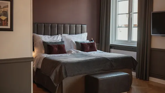 Viihtyisä hotellihuone, jossa on suuri sänky, lämpimän ruskeat seinät ja valoisa ikkuna. Sängyn jalkopäässä on mukava penkki.