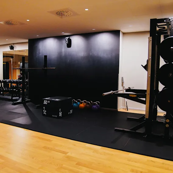 A gym area contains weights, benches, kettlebells, and exercise equipment on wooden and padded flooring, with a black wall and bright overhead lighting.