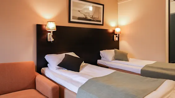 Two neatly made single beds with green throws against a dark wooden headboard, flanked by wall lamps. An orange armchair sits beside the beds in a warmly lit room.