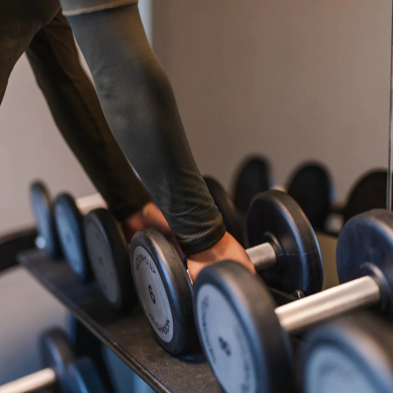 Dumbbells resting on a rack, hands gripping a six-kilogram weight. Mirror reflects the action in a gym setting.