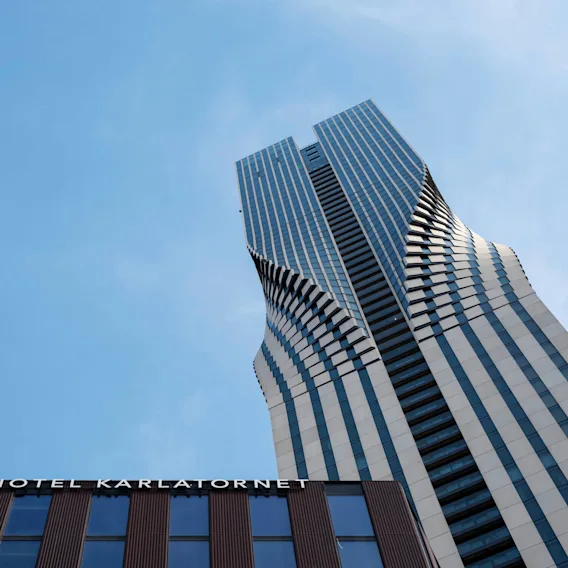 A skyscraper looms against a blue sky, its design features undulating patterns. "HOTEL KARLATORNET" is written on the lower building.