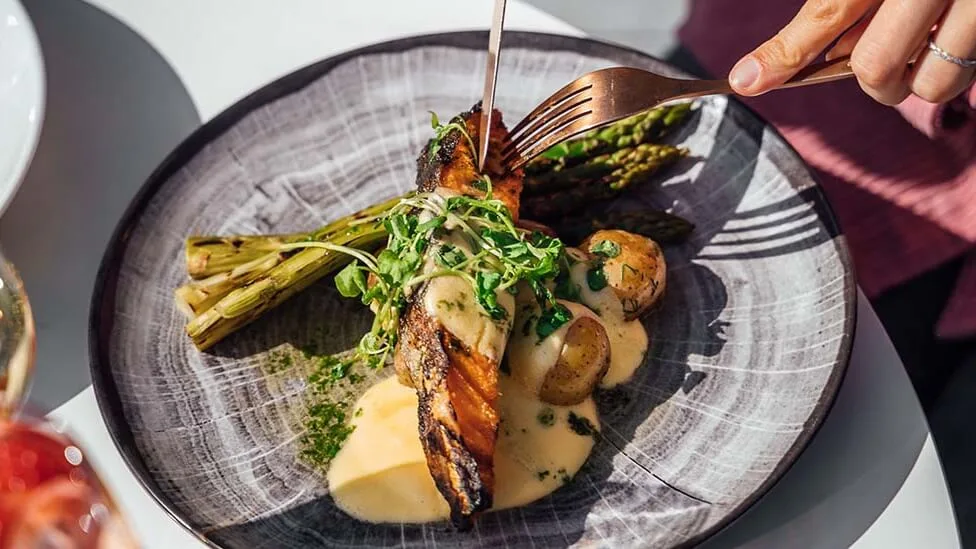 A plate of grilled salmon with asparagus, potatoes, and sauce; a person is cutting into the fish with a fork.