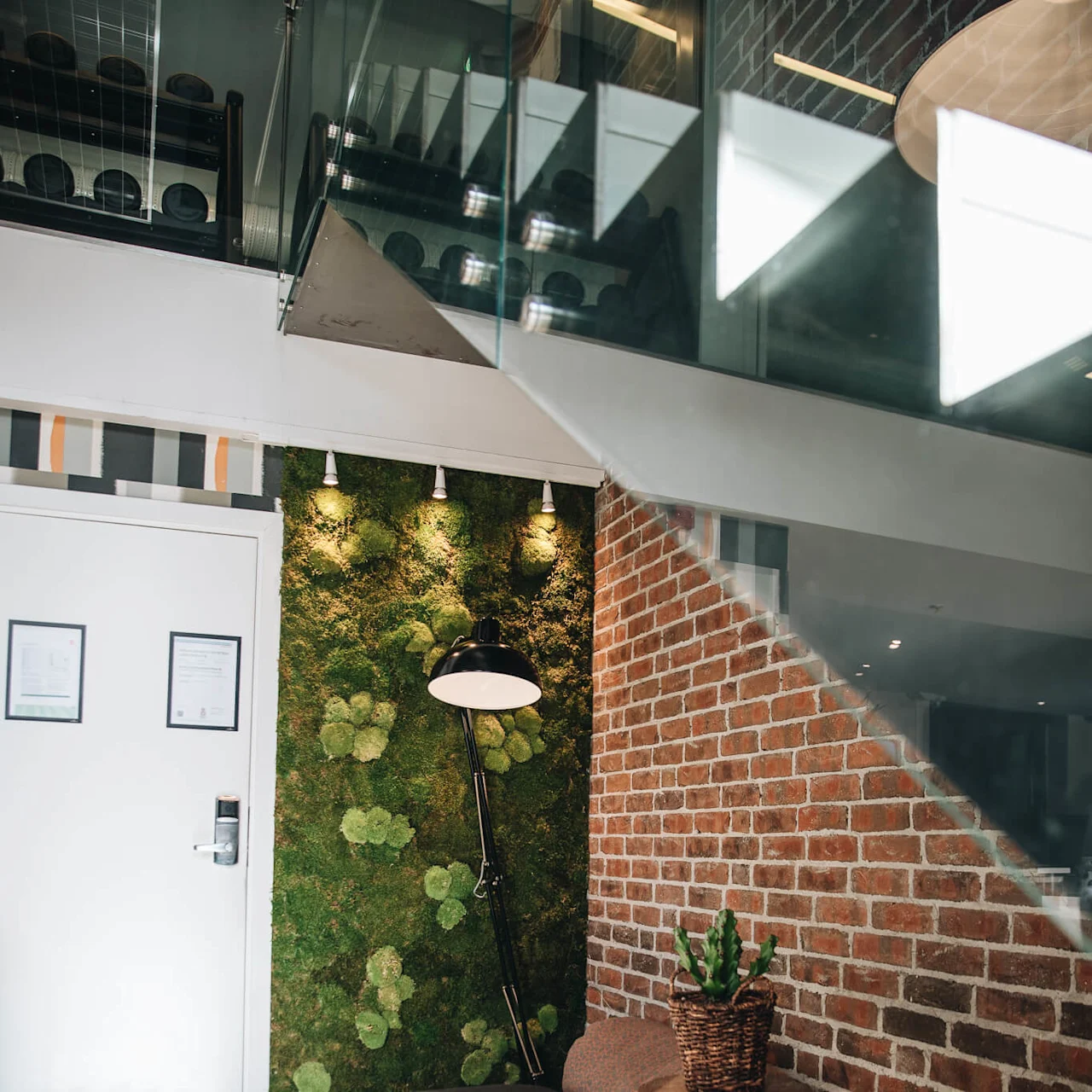 Large floor lamp stands by a moss-covered wall, near bean bags and a potted plant, in a cozy room with a glass staircase above and brick accents.