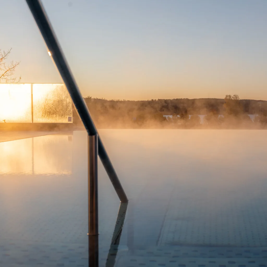 En dampende pool med et metalrækværk er placeret udendørs ved solnedgang, omgivet af træer og fjerne bakker under en klar himmel.