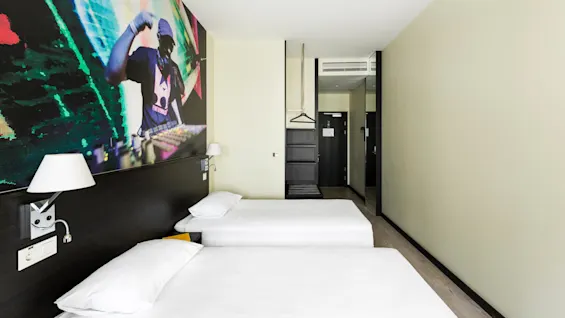 Två sängar med vita lakan är placerade i ett modernt hotellrum. En färgglad DJ-väggmålning finns på väggen, och en galge och hylla är nära ingången.
