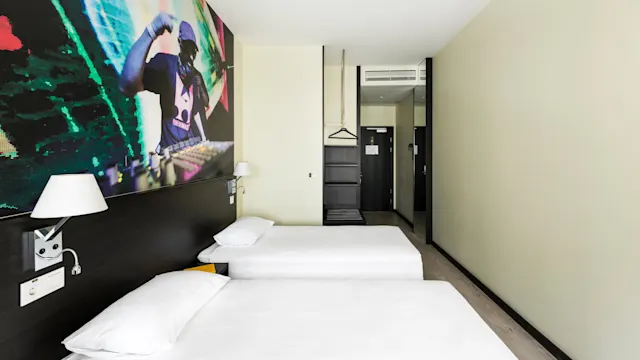 Two beds with white linens are located in a modern hotel room. A colorful DJ mural is on the wall, and a hanger and shelf are near the entrance.