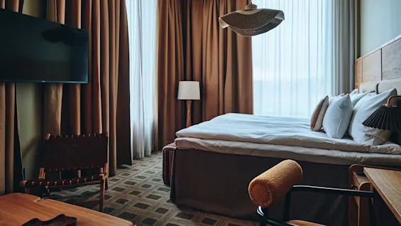 Et koselig hotellrom med en komfortabel seng, et stort vindu med gardiner, en veggmontert TV og stilige møbler.