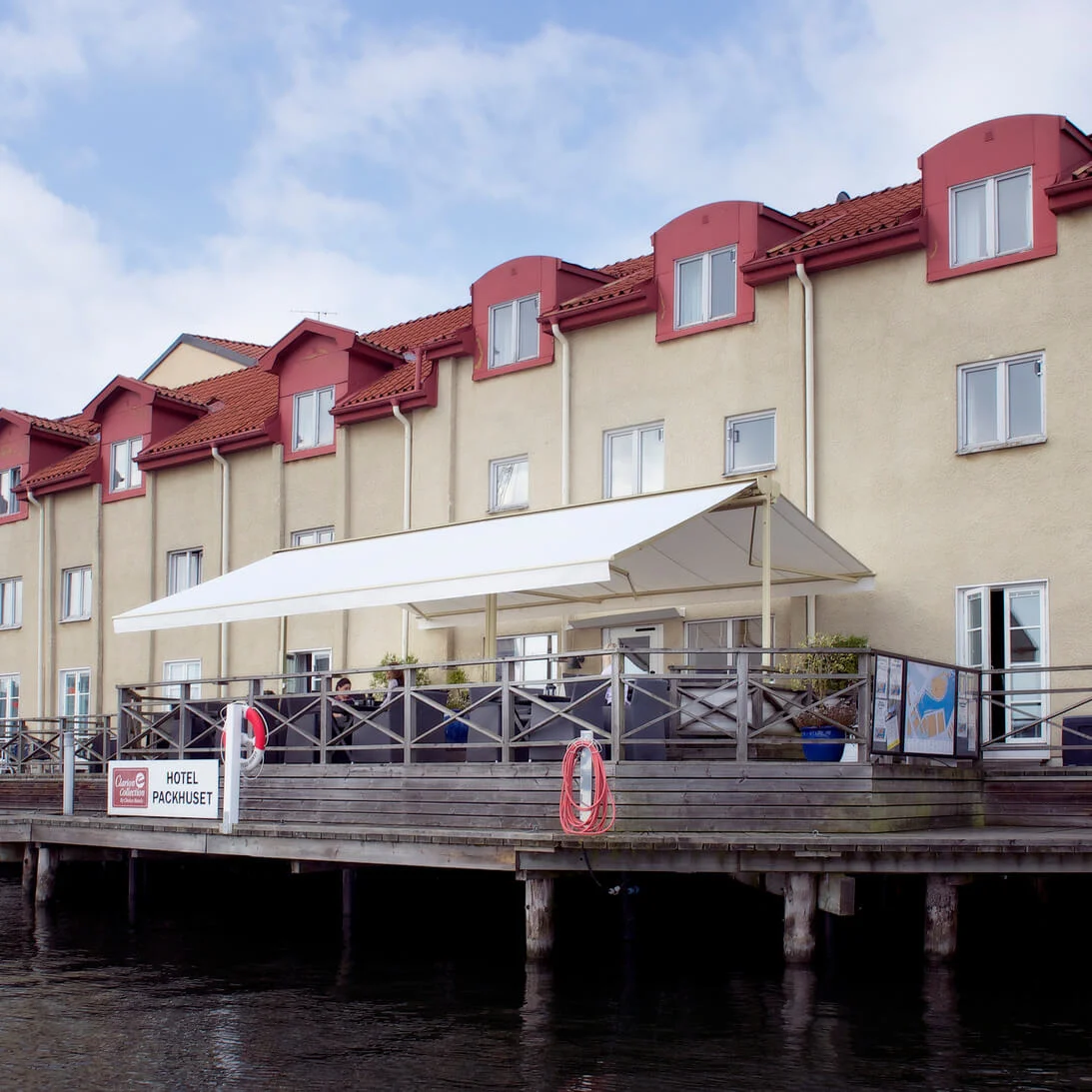 A waterfront building with a terrace extends over water, under a cloudy sky. Sign reads "HOTEL PAKKHUSET".
