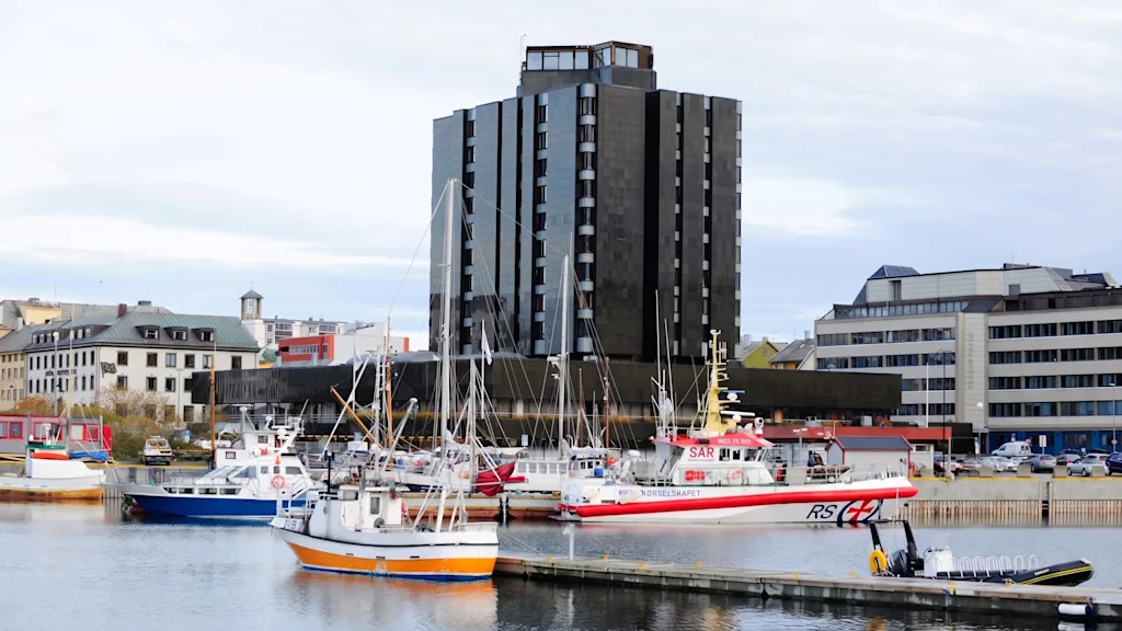 Et mørkt høybygg ved en havn fylt med båter under en overskyet himmel.