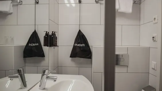 Sink with shiny faucet reflects in a bathroom mirror beside bags labeled “HAIRDRYER” and “HYGIENIA.” The white-tiled room includes towels on a rack, soap dispensers, and a tissue holder.