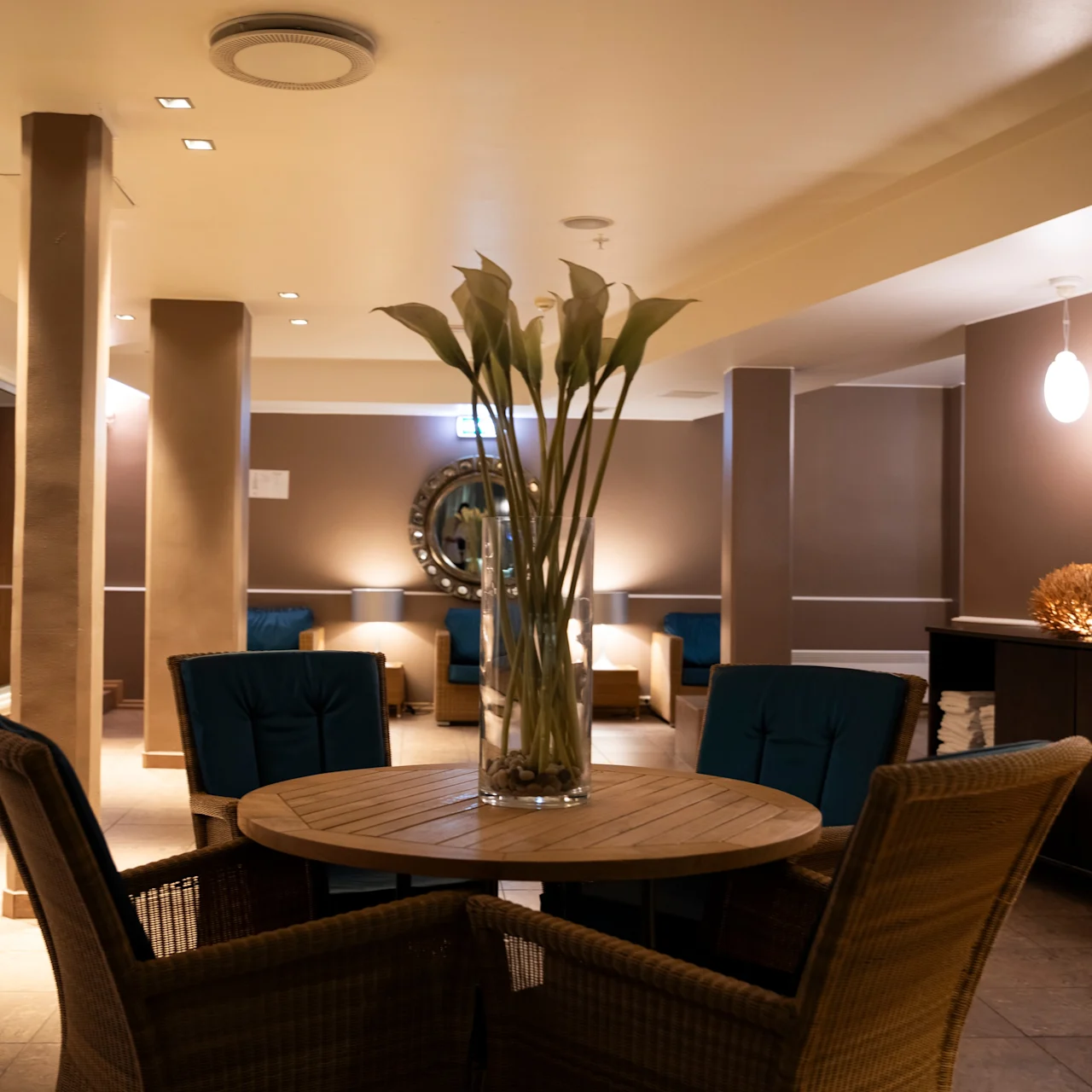 A round table with a vase of flowers is surrounded by wicker chairs in a dimly-lit, modern lounge. Soft lighting and decorative elements create a cozy atmosphere.